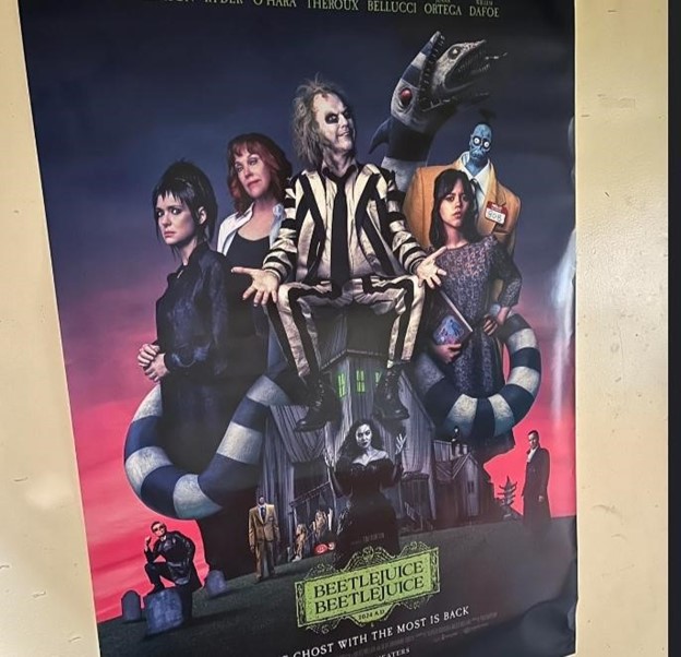 This is a popular poster at movie theaters for the new "Beetlejuice Beetlejuice." It features the main character Beetlejuice in the middle (Micheal Keaton) and to the direct left of him with light short hair is Barbra (Geena Davis). To the left of her is Lydia Deetz (Winona Ryder). And to the right of Beetlejuice is Asther (Jenna Ortega).  